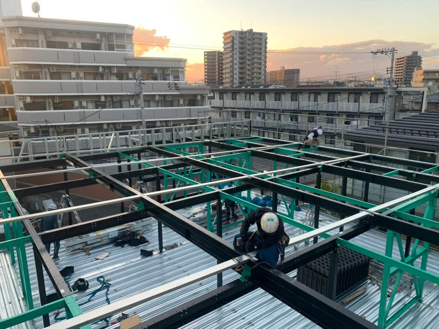 夕暮れの空のもと、鉄骨建方が進む建設現場で作業する職人たち