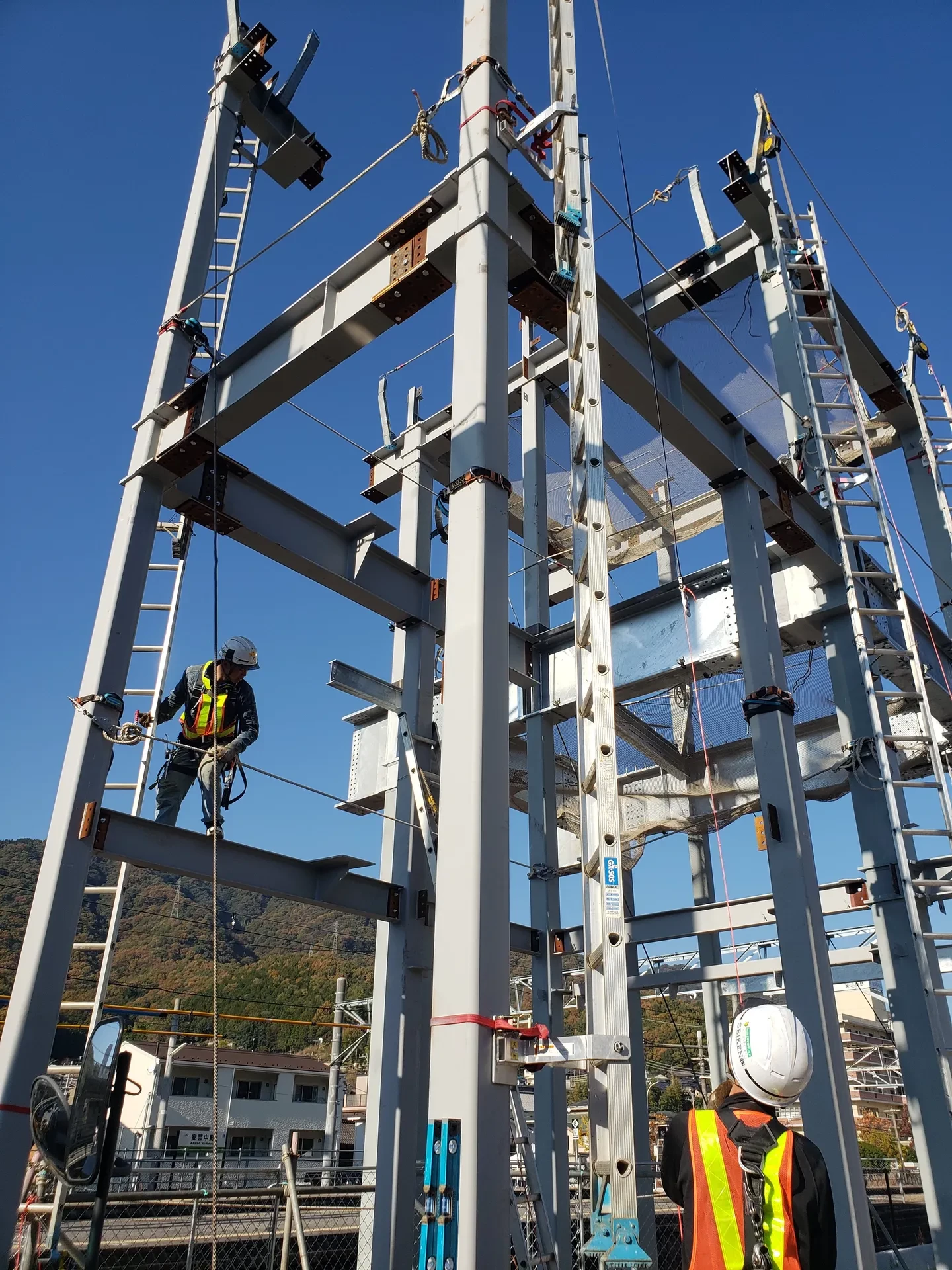 青空のもと、鉄骨建方の現場で梁の上を移動する職人